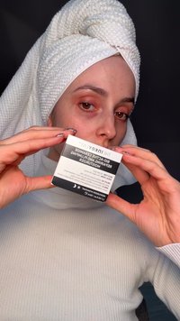 Personne avec une serviette blanche enroulée autour de la tête et un col roulé blanc tenant une boîte de produit de soin de la peau étiquetée pour une crème hydratante réparatrice et repulpante.