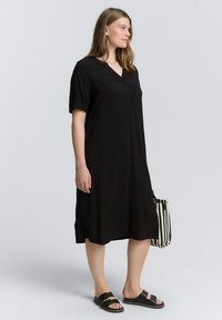 Vestido negro de manga corta y escote en V, con forma fluida y textura suave, combinado con sandalias negras y un bolso de rayas.