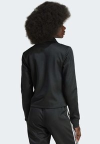 Jaqueta atlética preta com uma textura suave, apresentando um colar alto e três riscas brancas em ambas as mangas. Mostrada de costas.