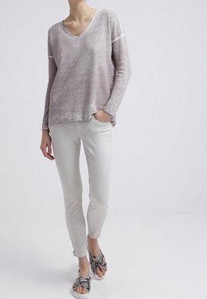 Femme portant un pull gris clair col en V, un pantalon blanc slim et des sandales ouvertes à motif peau de serpent, debout avec une main dans la poche.