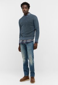 Niebieski sweter z dzianiny nałożony na koszulę w kratę, jasnoniebieskie dżinsy i brązowe buty sznurowane. Luźny strój z teksturowanych materiałów i warstwowym designem.