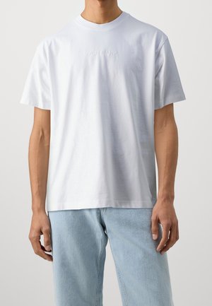 Uomo che indossa una semplice t-shirt bianca Guess Jeans e jeans azzurri, in piedi con le braccia rilassate lungo i fianchi su uno sfondo neutro.