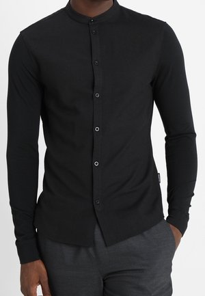 Muž nosící přiléhavou černou košili s mandlovým límcem, s knoflíky vpředu a tmavě šedé kalhoty.