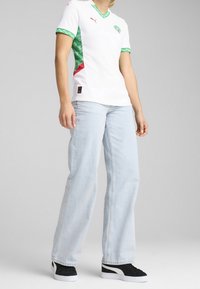 Persona vestida con una camiseta deportiva blanca y verde con un logo, vaqueros de pierna ancha azul claro y zapatillas negras y blancas, de pie sobre un fondo plano.