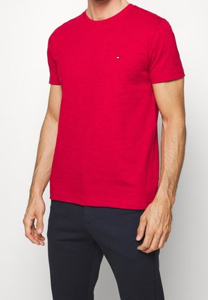 Mężczyzna w jasnoczerwonej, krótkiej koszulce z małym logo na lewej piersi i ciemnych spodniach na jednolitym tle.