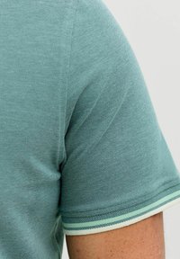 Τυρκουάζ πλεκτό πουκάμισο με κοντά μανίκια και μανσέτες με ρίγες σε ανοιχτό πράσινο και λευκό, φορεμένο στο χέρι ενός ατόμου σε απλό φόντο.