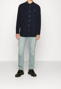 Granatowa koszula na guziki z długimi rękawami i kołnierzykiem, wykonana z teksturowanej tkaniny. Na piersi znajduje się małe żółte logo. Noszona z jasnymi jeansami i czarnymi butami.