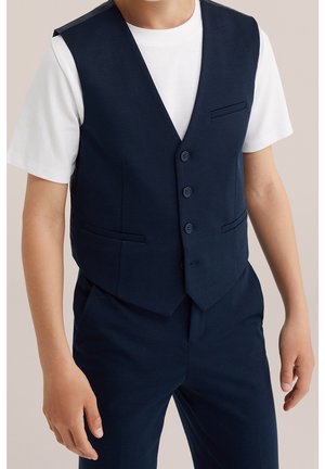 Ung person iført en mørkeblå knappet vest over en hvid kortærmet T-shirt og matchende mørkeblå bukser, stående med afslappede hænder.