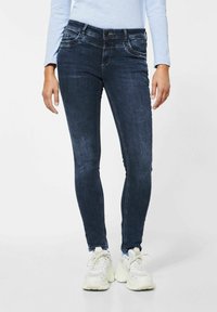 Mörkblå slim-fit jeans med subtila blekningar, framfickor och metalliska detaljer vid midjan. Matchas med vita sneakers.