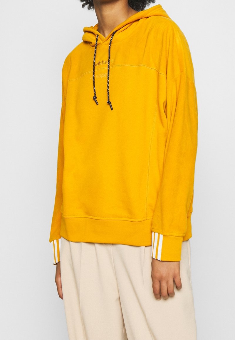 Sweatshirt à capuche jaune avec des cordons noirs, poignets côtelés ornés de trois rayures blanches et une coupe décontractée. Tissu doux et texturé.