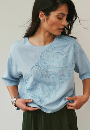 Femeie purtând o bluză bleu cu model texturat în spirale, fustă verde închis, cercei argintii și brățară, ajustând tivul bluzei.