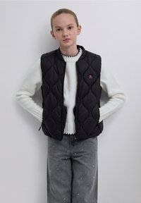 Musta tikattu liivi vetoketjulla ja pyöreällä violetilla logolla, päällä valkoinen neule, yhdistettynä harmaisiin farkkuihin, joissa koristeelliset yksityiskohdat.