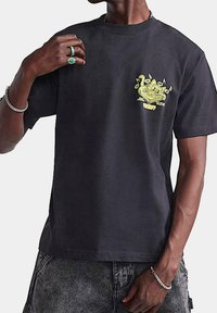 Camiseta de algodón negra con un diseño gráfico amarillo brillante en el lado izquierdo del pecho. Manga corta, cuello redondo y corte relajado.