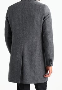 Hombre vistiendo un abrigo de lana gris visto desde atrás, con mangas largas y pantalones negros visibles debajo.