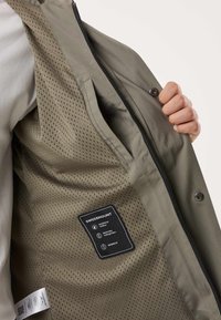 Ruka drži otvorenu zelenu jaknu koja pokazuje prozračni mrežasti podstava i crnu oznaku SWEDEMOUNT s detaljima o vodootpornosti i vjetrootpornosti.