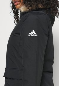 Frau trägt eine schwarze Adidas-Jacke mit einer mit Fell gefütterten Kapuze, das Logo ist auf dem oberen linken Ärmel zu sehen.