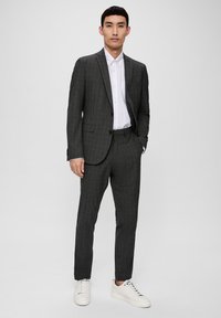 Costume gris charbon avec un motif à chevrons discret, coupe ajustée, veste à un seul bouton et pantalon assorti, associé à une chemise blanche et des baskets blanches.