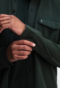 Dunkelgrünes Hemd mit Knöpfen und einer Brusttasche, sichtbaren schwarzen Knöpfen und hochgekrempelten Ärmeln, das eine weiche, glatte Stofftextur zeigt.