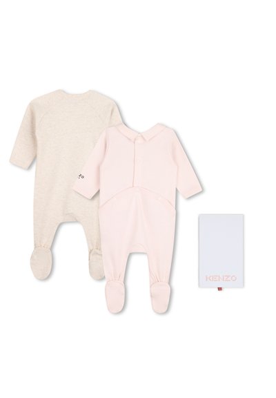 Deux pyjamas pour bébé à pieds pliés, l'un crème et l'autre rose pâle, à côté d'une étiquette ou carte blanche de la marque Kenzo.