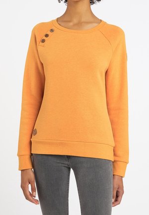 Kvinde iført en orange sweatshirt med lange ærmer og tre knapper nær venstre skulder samt tætsiddende grå jeans.