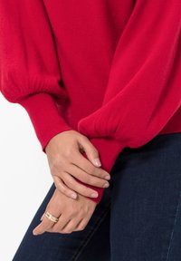 Maglione rosso lavorato a maglia con maniche a palloncino, polsini a costine e una texture morbida. Indossato con jeans blu scuri e un anello dorato alla mano.