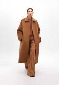 Dlouhý kabát se zapínáním vpředu vyrobený z měkké hnědé vlny s límečkem. Model má na sobě ladící hnědé kalhoty a blůzu na knoflíky.
