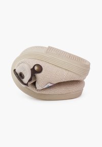 Zapato beige tipo slip-on con parte superior de tela texturizada y suela de goma. Presenta un diseño plegado con un botón metálico decorativo y acentos de costura.