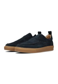 Zapatos de ante azul oscuro tipo slip-on con suela de goma color tostado, punta redondeada y un sutil detalle de costura en la parte superior. Presenta pequeñas pestañas en el talón.