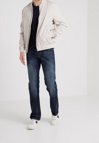 Světle béžová bomber bunda s žebrovanými manžetami a lemem, nošená přes námořnickou košili a v kombinaci s tmavě modrými džíny a bílými teniskami.