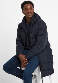 Smiling man wearing a navy long padded jacket, dark sweater, blue jeans, and white sneakers, sitting on a white stool.