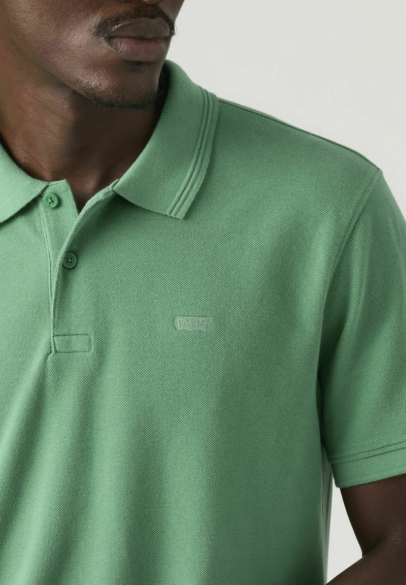 Hombre con camisa polo verde de Levi's, mostrando el cuello, los botones y un pequeño logo en el pecho, sobre un fondo liso.