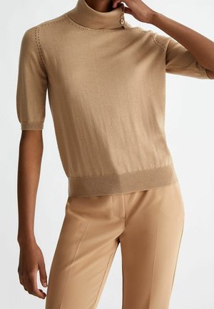 Mujer con jersey beige de cuello alto y mangas cortas con detalles de botones y pantalones beige de talle alto a juego, sobre un fondo liso.