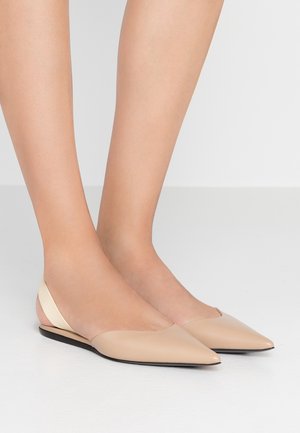 Fødder iført beige spidse slingback-flats med sort sål på en lys gulvbaggrund.