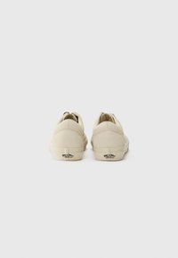 Beige canvas low-top sneakers with a rubber sole, featuring a black accent tag on the heel, smooth texture, and rounded toe design.