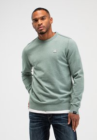 Hellgrüner Baumwollpullover mit langen Ärmeln, gerippten Bündchen und Saum sowie einem kleinen Logopatch auf der Brust. Getragen über einem weißen Hemd und Jeans.