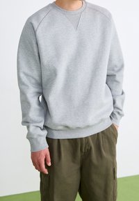 Grauer Sweatshirt mit rundem Halsausschnitt und Rippenbündchen, das eine subtile Textur aufweist. Getragen über olivfarbenen Cargo-Hosen vor einem schlichten Hintergrund.