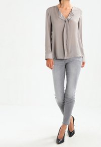 Blusa di seta grigia con scollatura a V e dettaglio con nodo, maniche lunghe, abbinata a jeans skinny grigi chiari e tacchi alti neri. Design minimalista.