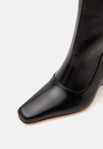 Bottine en cuir noir brillant avec bout carré et finition lisse, présentée sur un fond clair uni.