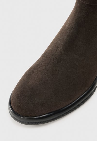 Bottine en daim marron, à bout rond, texture lisse, semelle en caoutchouc noir avec une légère bordure, détails de couture minimaux.