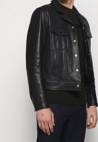 Uomo che indossa una giacca di pelle nera con tasche a pattina abbottonate sopra una camicia nera e pantaloni scuri, mostrato dalle spalle ai fianchi.