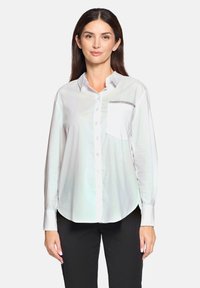White button-up shirt in lightweight fabric with a chest pocket featuring a black trim accent. Long sleeves, curved hem, and smooth texture.