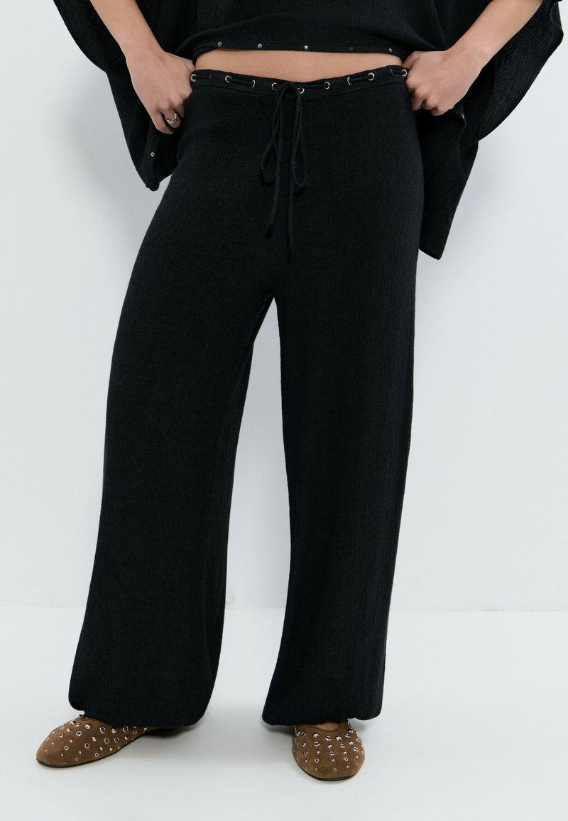 Pantalones de punto negros de pierna ancha con cintura ajustable con cordón y zapatos marrones sin cordones con detalles de ojales plateados, vistos desde la cintura hacia abajo.