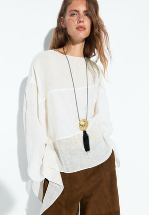 Frau mit welligem Haar trägt eine lockere weiße Bluse, braune Hose und eine lange Kette mit goldenem Anhänger und schwarzem Quast und steht vor einem schlichten Hintergrund.
