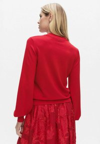 Czerwony dziany sweter z wysokim kołnierzem i długimi bufkowatymi rękawami. Posiada dekoracyjny guzik z tyłu oraz ściągacz na dole. Noszony na czerwonej kwiecistej spódnicy.