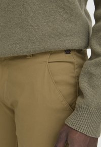 Suéter de punto verde oliva con puños acanalados, combinado con pantalones de algodón beige que cuentan con una pequeña etiqueta de logo y bolsillos laterales.