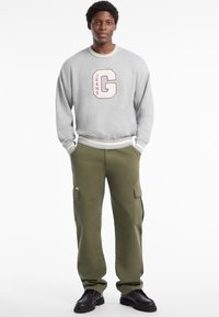 Felpa grigia con ricamo maroon della lettera "G" e "JEANS", polsini e orlo a coste. Pantaloni cargo oliva con molteplici tasche. Calzature nere.