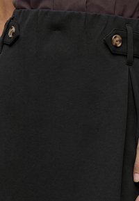 Gros plan sur la taille d'une jupe noire avec deux boutons marron sur les bretelles et une partie d'une main reposant à côté.