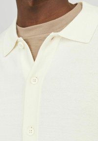 Kreemivärvi polo särk, mis on valmistatud tekstuuriga kangast. Omab kraega kaelust ja kahte nööpi sulgemiseks. Kihtide kaupa beež särgi peal.