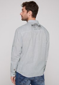Camisa de manga larga azul claro con un patrón texturizado, puños de botón y un logo bordado en la parte trasera. Combinada con jeans de mezclilla.
