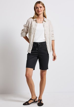 Shorts noirs avec une taille élastique à cordon, associés à un débardeur blanc et une chemise en lin beige clair à boutons. Sandales noires présentes.